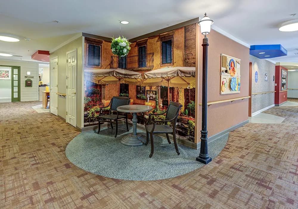 A hallway with a mural of a café scene, featuring two metal chairs, a small round table, and a streetlamp, creating an indoor seating area.