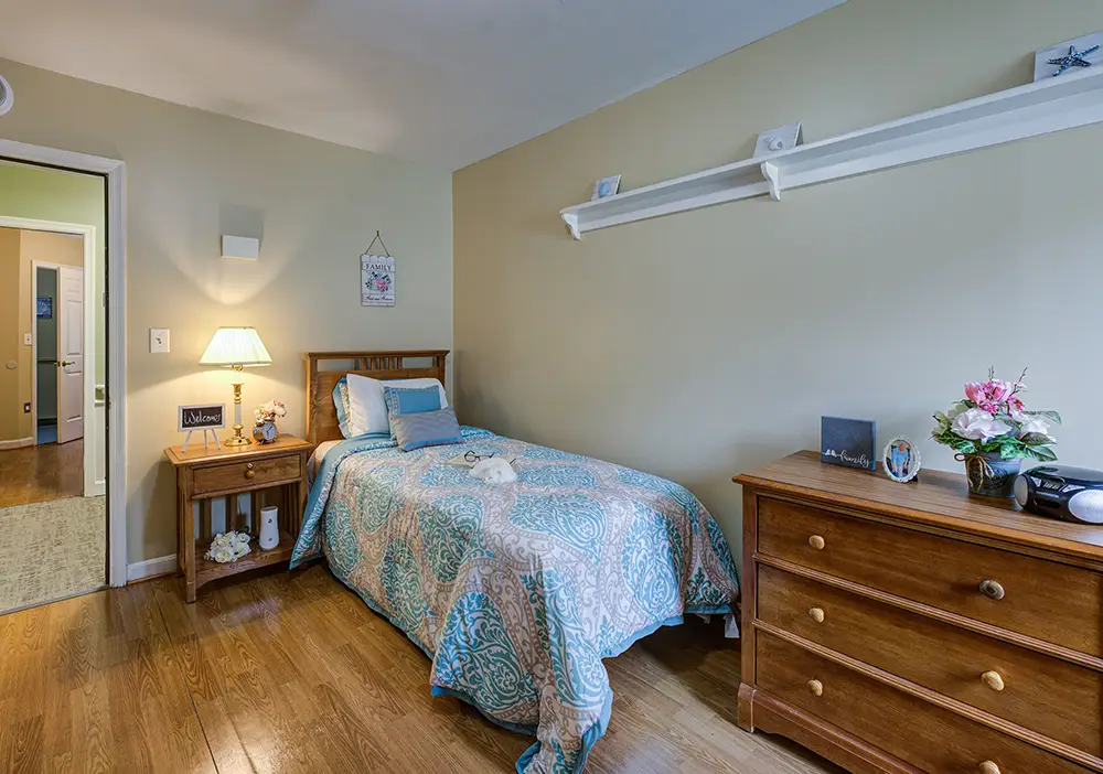 A neatly made single bed with a blue patterned quilt, wooden nightstand with a lamp, and a dresser in a tidy bedroom with hardwood flooring.