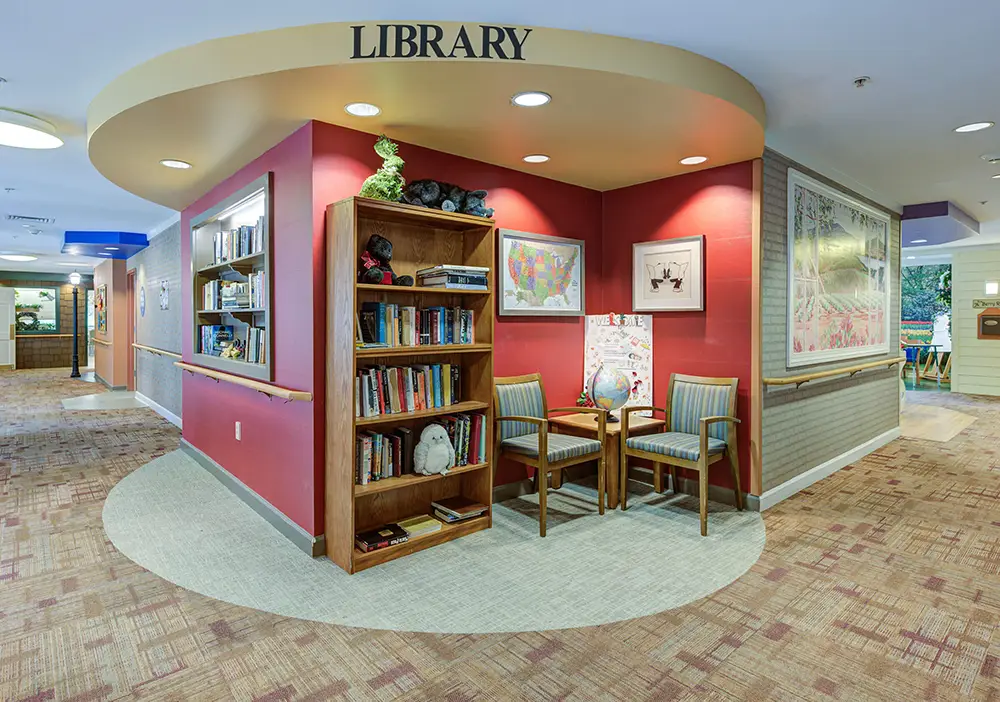 Library nook with built-in bookshelves, two chairs, a small table, and wall art in a well-lit hallway with carpeted floors.