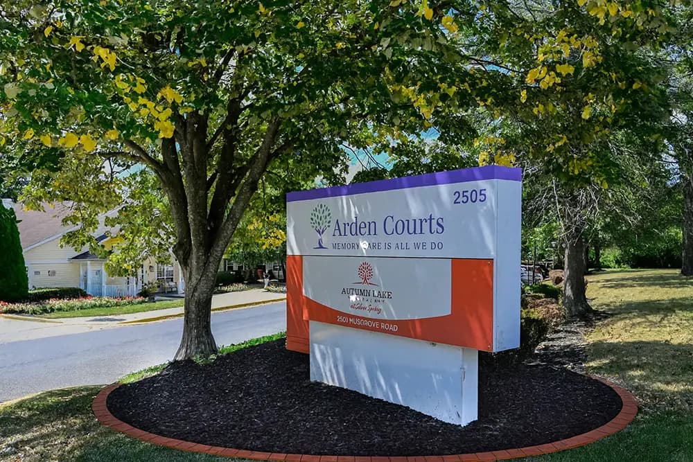 A large sign under a tree reads "Arden Courts, Memory Care is All We Do, 2505" and "Autumn Lake Healthcare," located on a landscaped area beside a road.